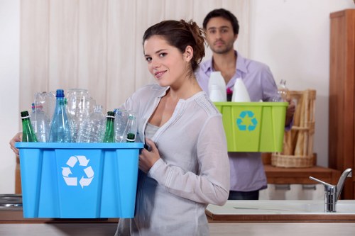 A young woman with dark hair styled in an updo is standing in a kitchen, smiling slightly while holding a blue recycling bin filled with clear glass bottles of various shapes and sizes. She is wearing a white long-sleeved top and appears to be preparing to recycle the bottles. Behind her, a man with dark hair and a beard is holding a green recycling bin with a recycling symbol, containing additional bottles and containers, standing near a wooden dish rack on the kitchen counter. The background includes a modern kitchen setting with a stainless steel sink, a faucet, and natural light illuminating the scene. This setting suggests a domestic environment focused on waste separation and recycling activities, in line with waste management services such as those provided by Commercial Waste Hoxton around the London area.