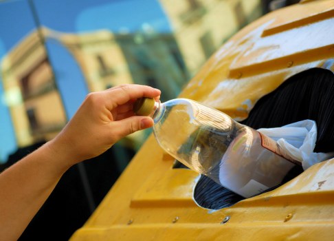 A person's hand is shown placing a clear, empty plastic bottle into a yellow waste disposal slot on a skip or receptacle, which is situated outdoors in a residential or commercial setting. The bottle appears to be a standard PET container with a plastic cap. The skip or bin has a wooden or painted surface with a smooth finish and round openings, designed for waste collection. In the background, a blurred view of buildings and trees can be seen, suggesting an urban or suburban environment, possibly within Hoxton or a similar area in London. The lighting indicates natural daylight, highlighting the clean appearance of the plastic bottle and the bright yellow color of the waste container. This scene reflects a waste disposal activity associated with rubbish removal services provided by Commercial Waste Hoxton, emphasizing recycling or waste separation practices in line with local rubbish collection standards.