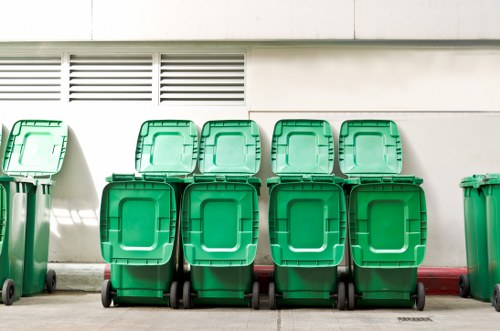 A row of eight green wheelie bins arranged in front of a white building with horizontal vented panels at the upper section. The bins are made of plastic with textured surfaces, and each has a hinged lid. Four bins are positioned directly on the pavement, with the remaining four slightly behind and resting against the first row. The pavement appears to be concrete or similar material, and a red line runs along the base of the building wall. The environment suggests an outdoor commercial or residential waste collection area, with adequate space for rubbish removal services. The image conveys a clean, organized waste disposal setup, characteristic of services provided by companies like Commercial Waste Hoxton in the Hoxton area.