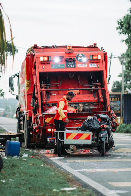 A small street scene showing a mobile rubbish cart loaded with various waste materials, including cardboard boxes, black plastic garbage bags, and other debris. The cart is positioned in the foregroun