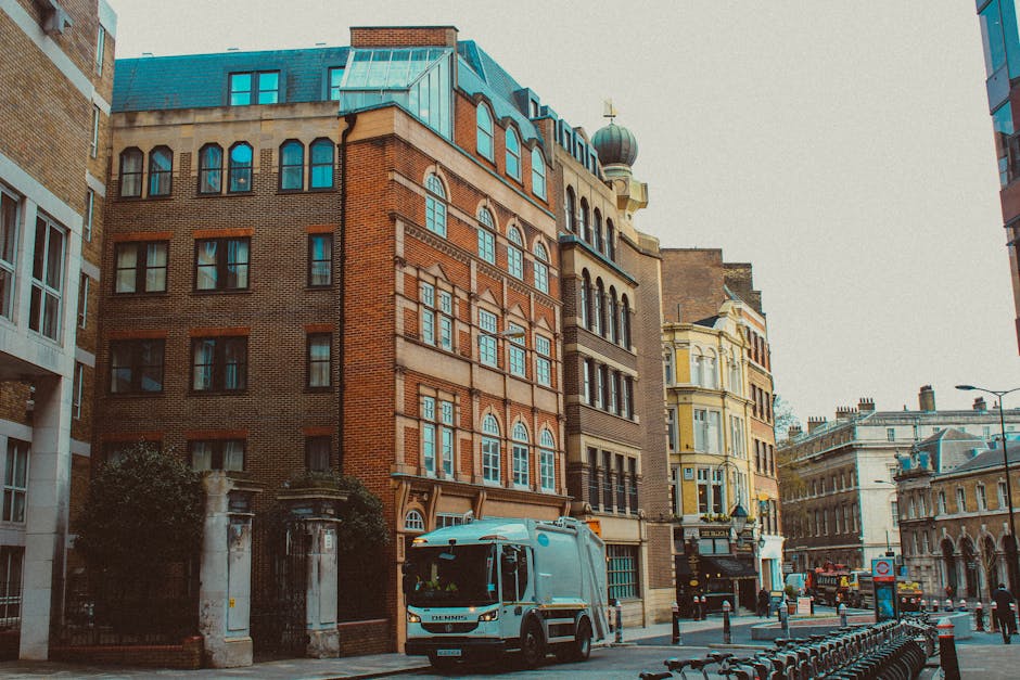 The image depicts a city street scene with a row of multi-story buildings featuring a mix of architectural styles, including red brick facades, ornate window frames, and various finishes. In the foreg